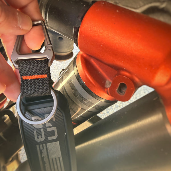Close-up of a key fob with a strap attached, showing brand logo raptor code orange.